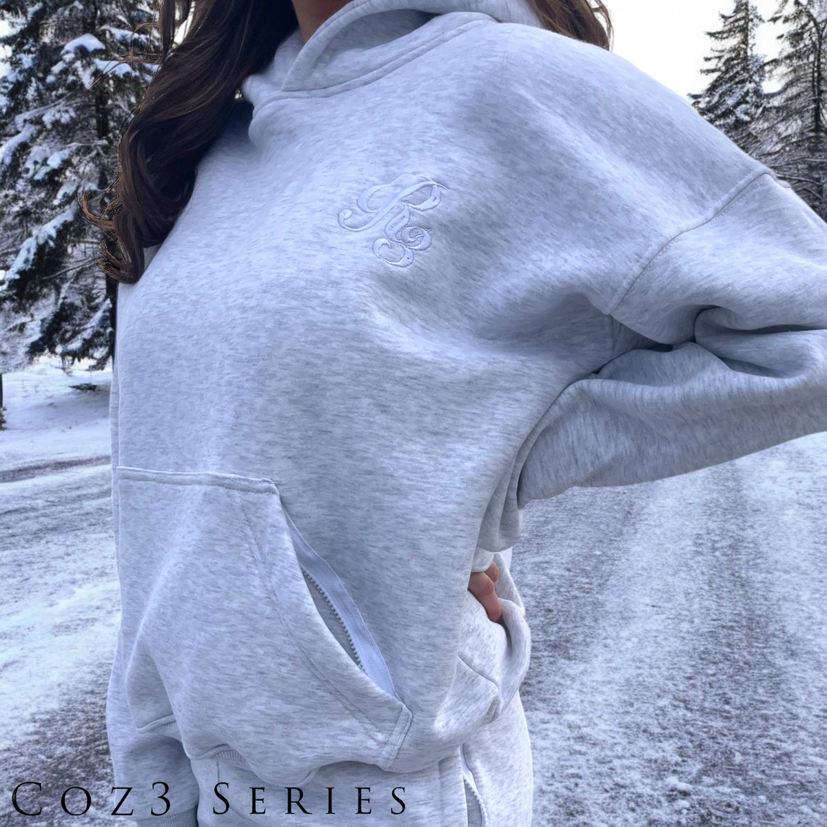 A person wearing a gray Coz3 Max Hoodie with the hood up, standing in a snowy outdoor setting.