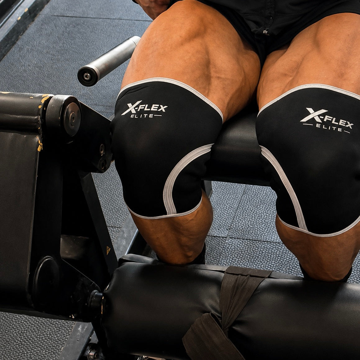 Close-up of a person's legs in a gym setting, wearing black X-FLEX ELITE knee sleeves with white trim while performing a leg extension exercise.