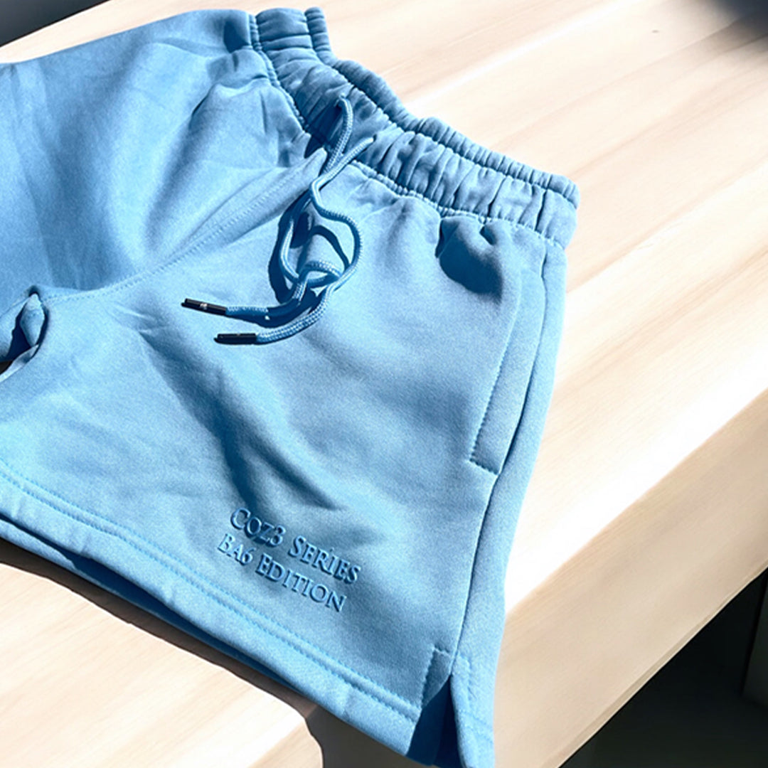 An array of Coz3 Ba6 Edition Shorts in various colors including pink, blue, purple, white, and gray, displayed in a line with a soft focus background. The shorts are characterized by a relaxed fit, 3-inch waistband, and drawstrings.