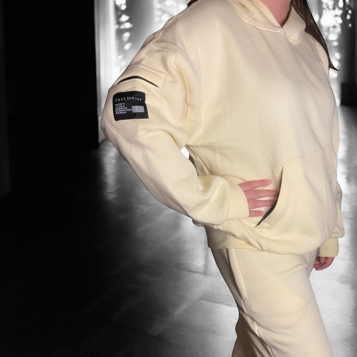 A person wearing a butter cream Coz3 Max Hoodie with the hood up, standing in a snowy outdoor setting.