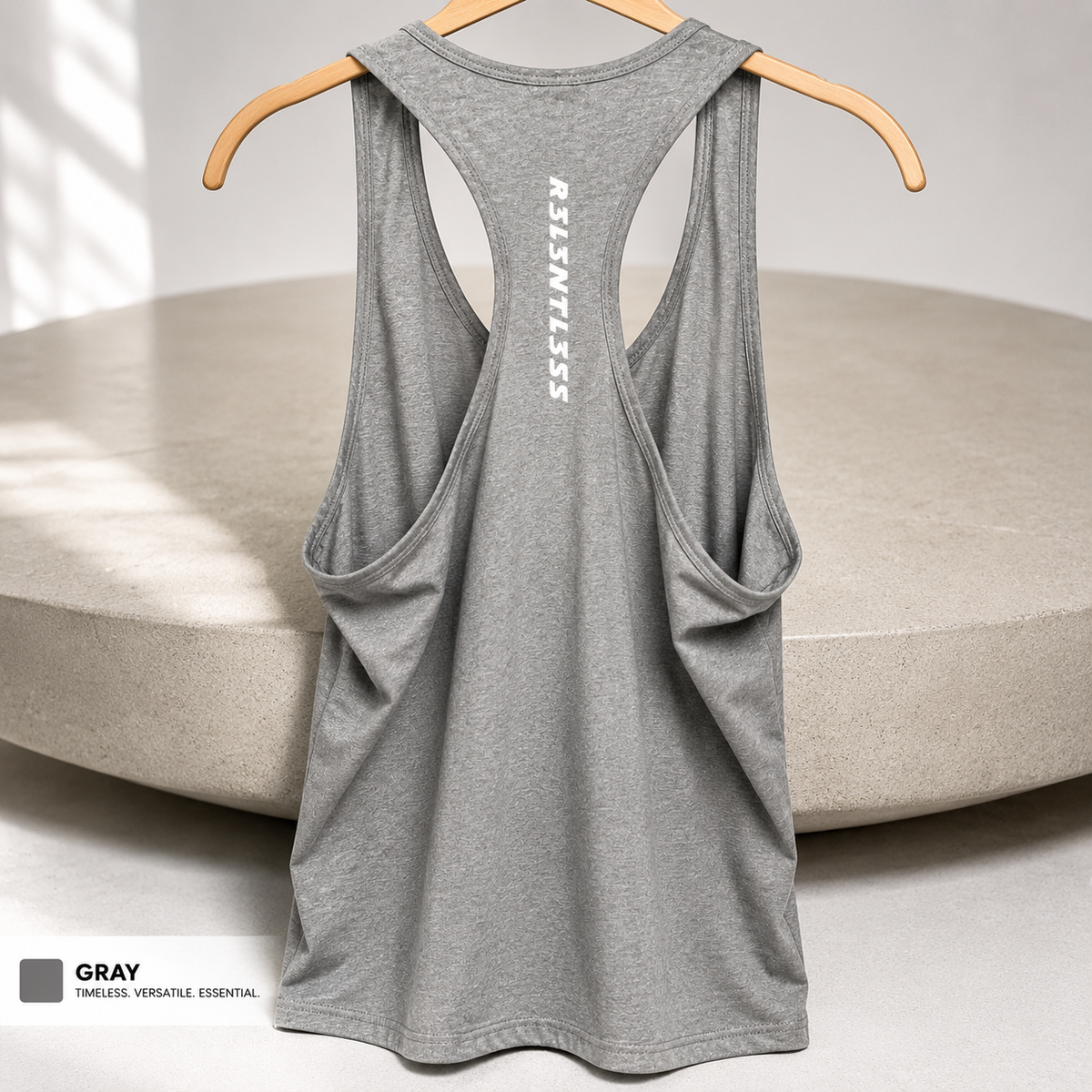 A heather gray racerback tank top hanging on a wooden hanger against a neutral studio backdrop with a circular platform behind it. The back of the tank features a vertical white 'RESTLESS' logo and side pockets.
