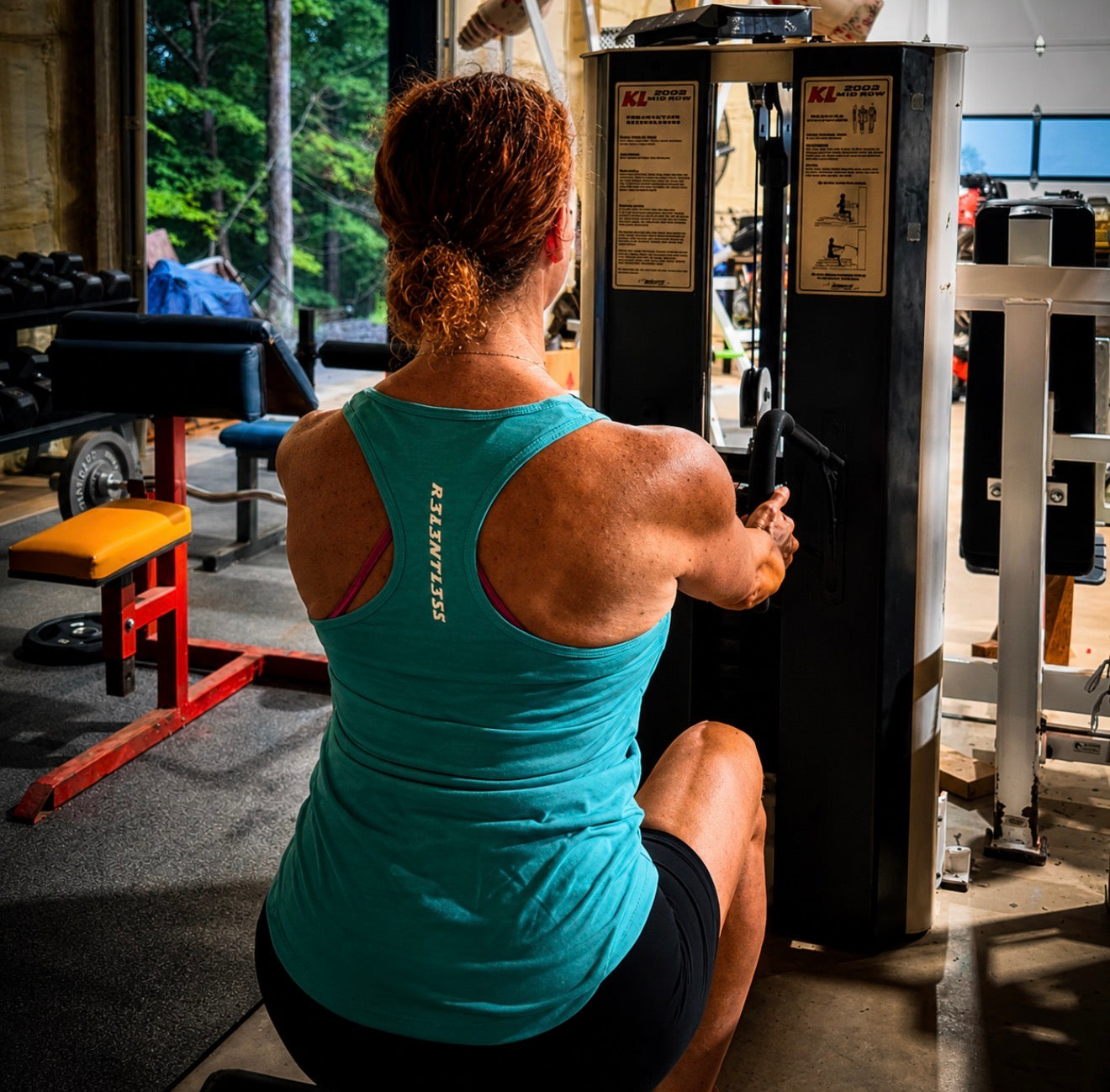 A woman in a teal ribbed tank top and black shorts is seen from behind, pulling a weight machine in a gym. The brand name 'REDEFINE' is printed vertically on the back of her tank top.