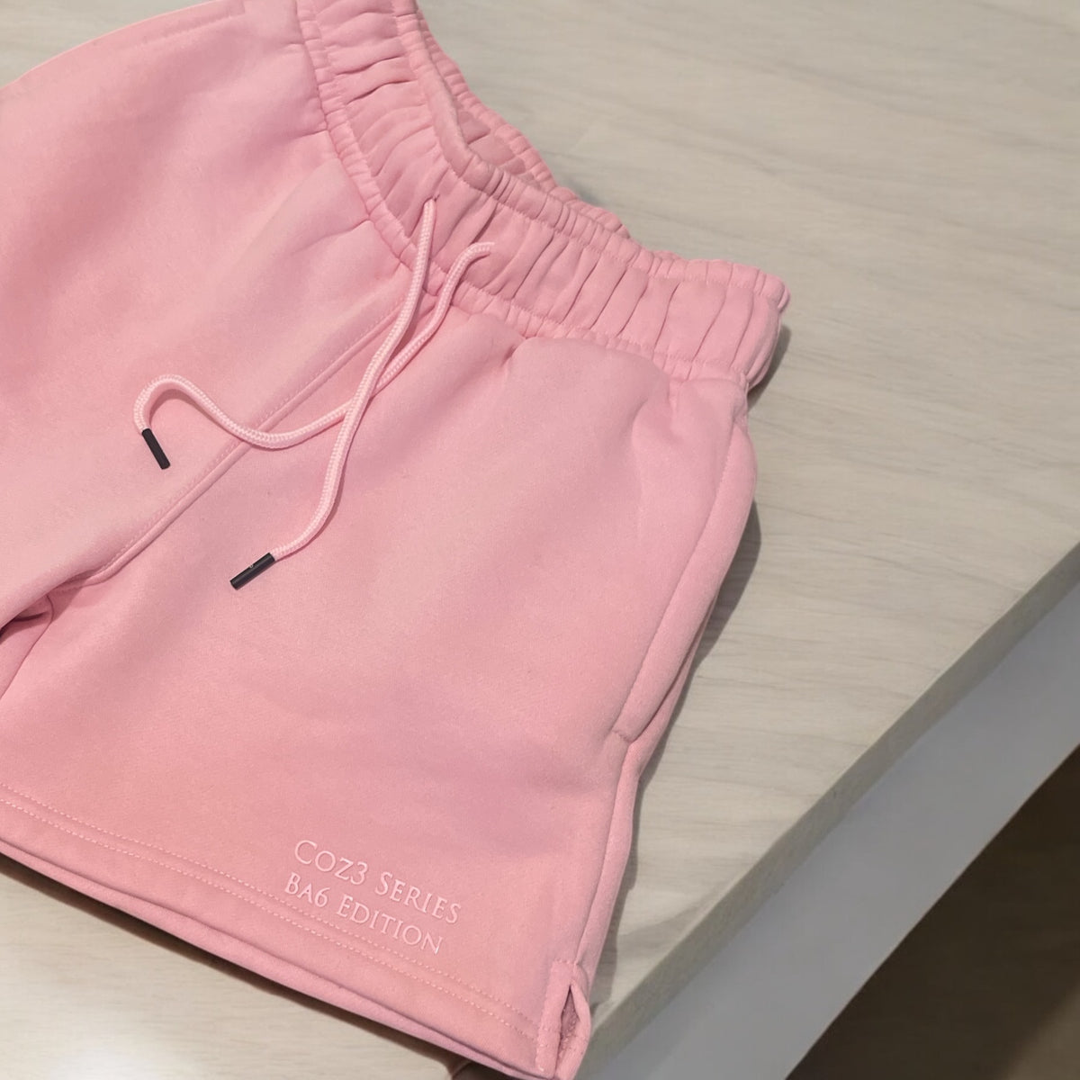 An array of Coz3 Ba6 Edition Shorts in various colors including pink, blue, purple, white, and gray, displayed in a line with a soft focus background. The shorts are characterized by a relaxed fit, 3-inch waistband, and drawstrings.