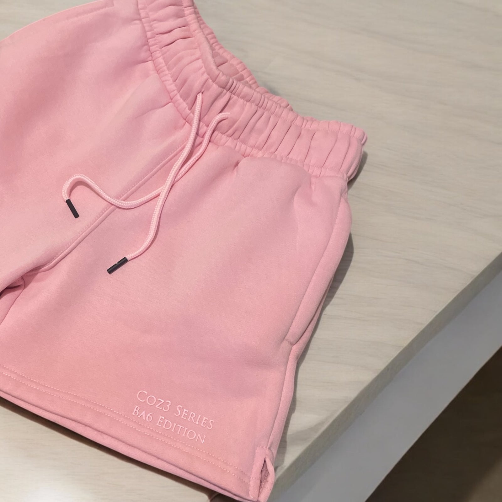 An array of Coz3 Ba6 Edition Shorts in various colors including pink, blue, purple, white, and gray, displayed in a line with a soft focus background. The shorts are characterized by a relaxed fit, 3-inch waistband, and drawstrings.